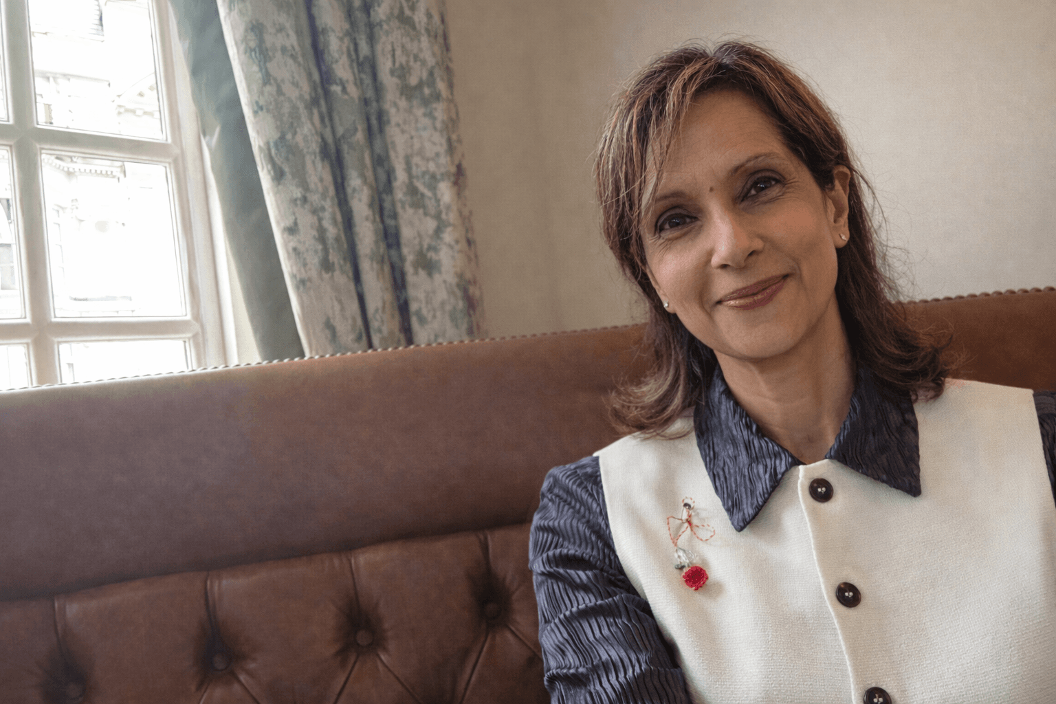Smiling woman with brown hair wearing a white vest, sitting on a tufted leather couch.